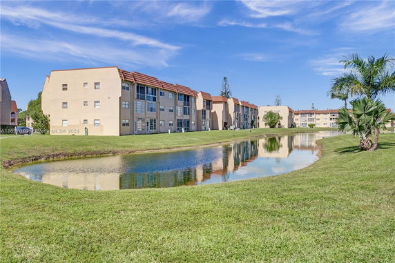 Three-story residential buildings viewed from the side with a pond and grassy area in front.