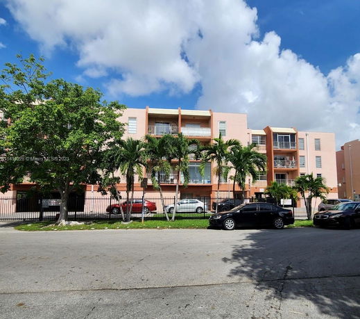 Front view of a multi-story apartment building with balconies and a fenced parking area.
