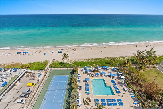 Panoramic view of a beachside area with a swimming pool, tennis court, and ocean in the background.