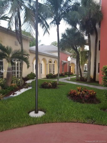 Front view of a building with arched windows and a light-colored roof, surrounded by palm trees and landscaped greenery.