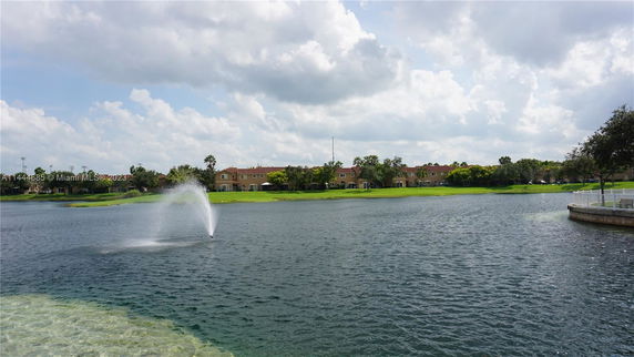 View of a lake with a fountain and houses in the background on a cloudy day.