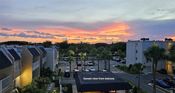 Panoramic view of sunset from a building balcony, overlooking parking area and surrounding buildings.