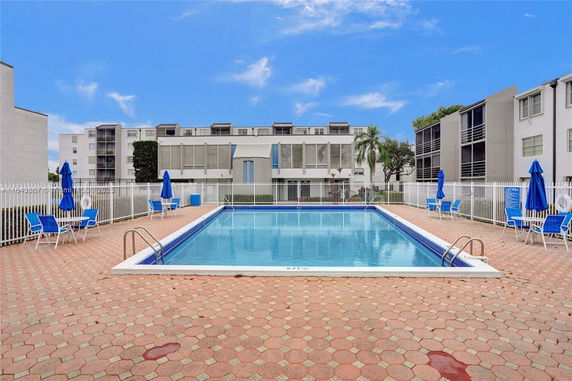 Outdoor swimming pool area with lounge chairs and umbrellas near apartment buildings.