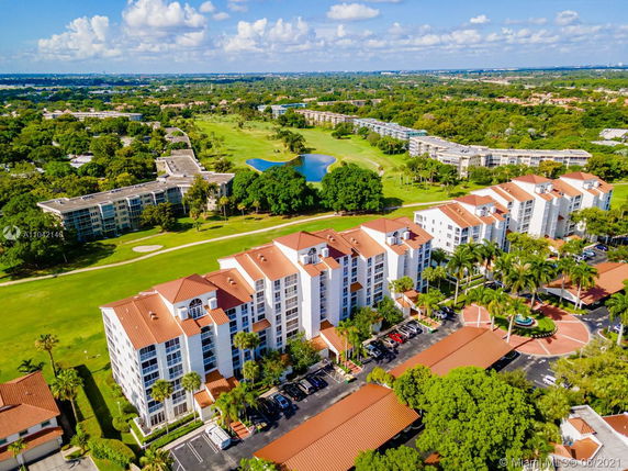 Aerial view of a residential complex with multiple multi-story buildings featuring terracotta roofs, surrounded by green lawns and trees, with a golf course in the background.