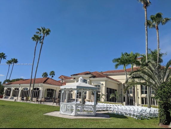 View of a large building with a red-tiled roof and tall windows, surrounded by palm trees and a grassy area with chairs arranged in rows.