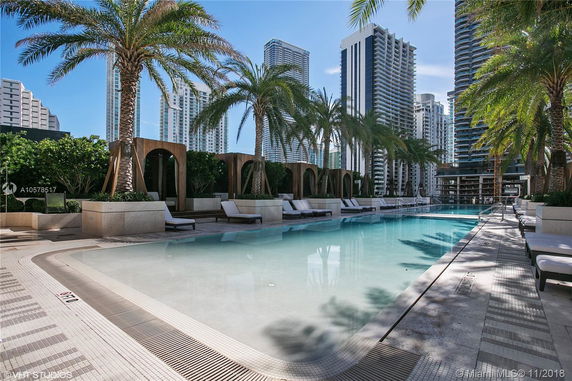 Rooftop pool area with lounge chairs and palm trees, surrounded by tall buildings.