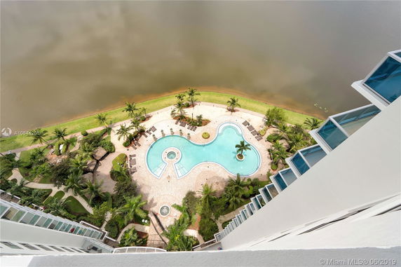 View from a high building overlooking a uniquely shaped swimming pool and landscaped area below, adjacent to a body of water.