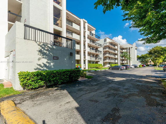 Front view of a multi-story apartment building with balconies and surrounding parking area.