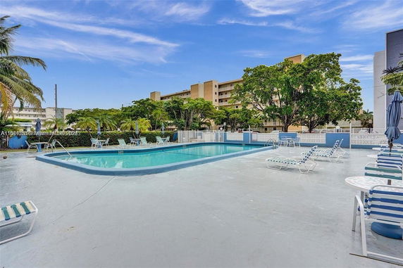 Outdoor swimming pool area with lounge chairs, surrounded by trees and nearby buildings.