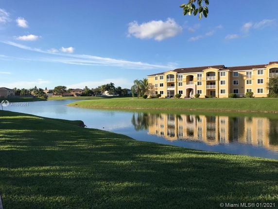 View of a three-story apartment building with a lake and green lawn in the foreground.