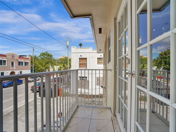View from a balcony overlooking a street with buildings and parked cars, featuring a railing and glass door.