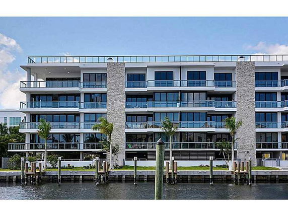 Front view of a multi-story modern building with large glass windows and balconies, overlooking a body of water.