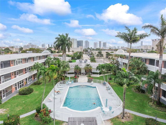 Aerial view of a residential complex with a central swimming pool surrounded by buildings and palm trees, with cityscape in the background.