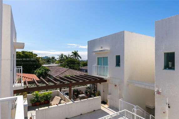 View from a building showing a white modern structure with small balconies and a patio area with outdoor furniture, surrounded by trees and palm trees in the background.