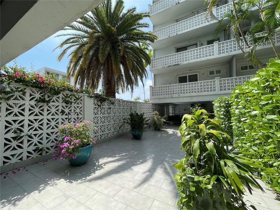 View of a multi-story building with latticework balconies, surrounded by plants and palm trees.
