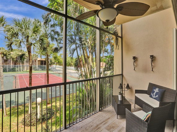 View of a tennis court from a balcony with outdoor seating and ceiling fan.
