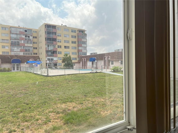 View from a window showing a multi-story building and a swimming pool area surrounded by grass.