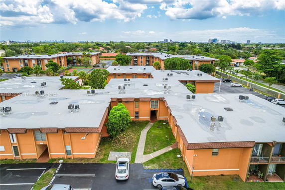 Aerial view of an apartment complex with several buildings and parking areas, featuring flat rooftops and surrounding greenery.