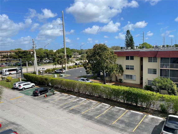 View of a multi-story residential building with parking area in front. Several cars are parked, and the street and other buildings are visible in the background.