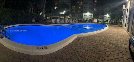 Night view of an illuminated swimming pool surrounded by deck chairs and adjacent to a building.