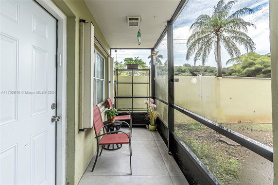 View from a narrow patio area outside a building with chairs and potted plants, featuring a white door and a window.
