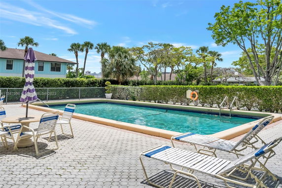 Swimming pool area with lounge chairs, an umbrella, and surrounding greenery, adjacent to a residential building.