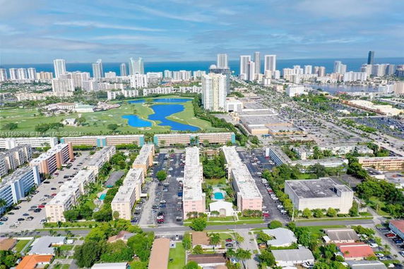 Aerial panoramic view of a cityscape with multiple residential and commercial buildings, a golf course, and high-rise structures in the background near the coastline.