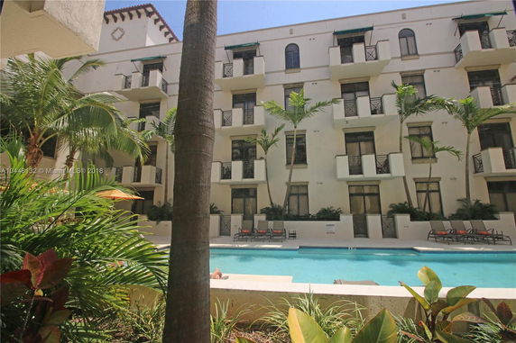 View of a multi-story building facade with balconies and a pool surrounded by palm trees.