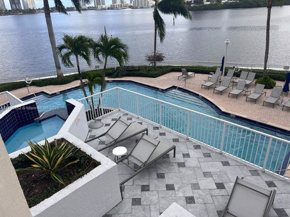 Outdoor view of a swimming pool area adjacent to a water body, surrounded by palm trees and lounge chairs.