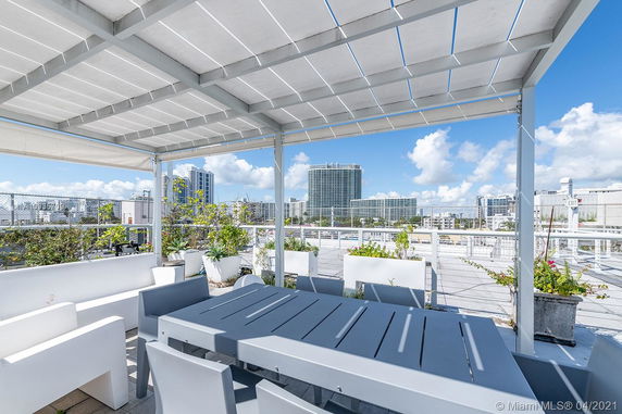 Rooftop area with seating and partial covering, surrounded by plants and overlooking city skyline.