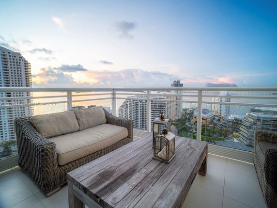 Balcony with a wicker sofa, wooden table, and view of city buildings and water.