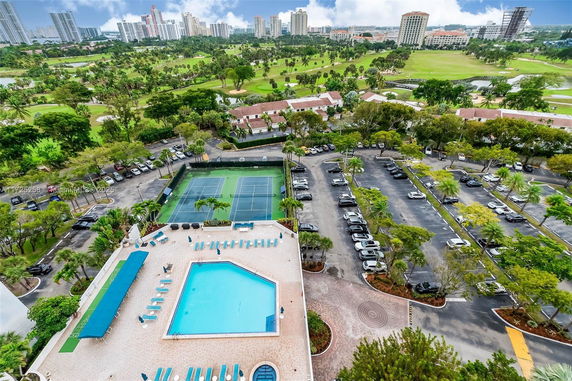 Panoramic view showing a swimming pool, tennis courts, parking area, and distant skyline.