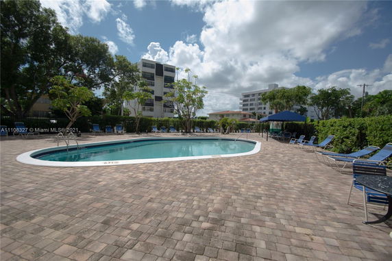 View of a swimming pool area surrounded by lounge chairs and nearby buildings.
