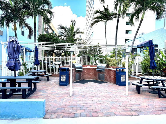 Outdoor area with picnic tables and brick barbecue grills, surrounded by palm trees and nearby buildings.