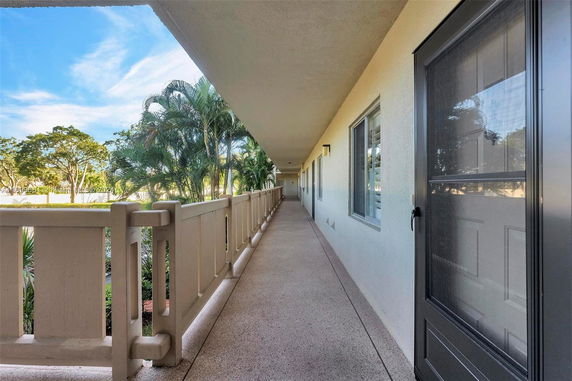 View along an outdoor corridor with railings and windows on one side, and trees visible beyond the railings.