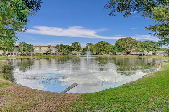 View across a pond towards buildings and a fountain, surrounded by trees.