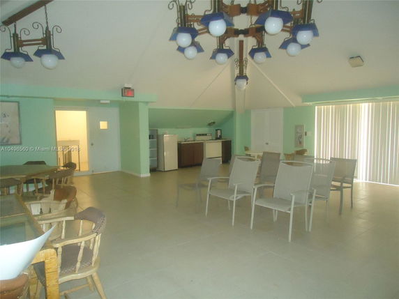 Room with chairs and tables, light fixtures hanging from the ceiling, and a small kitchenette in the corner.