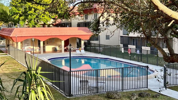 Outdoor swimming pool surrounded by a fence and a small shaded area with chairs.