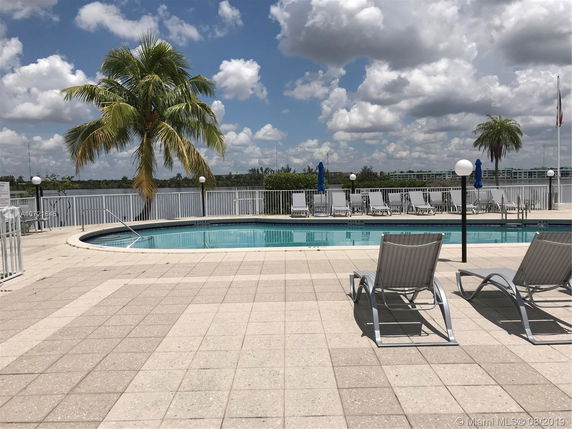Outdoor swimming pool area with lounge chairs, palm trees, and a view of the water and distant buildings.