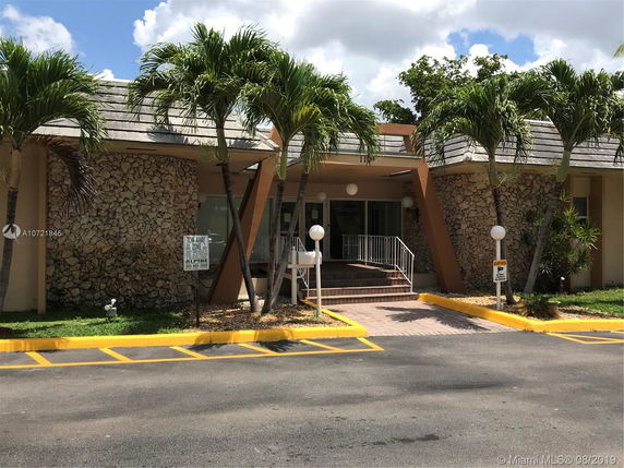 Front view of a building with a stone facade and sloped roof, featuring palm trees and stairs leading to the entrance.