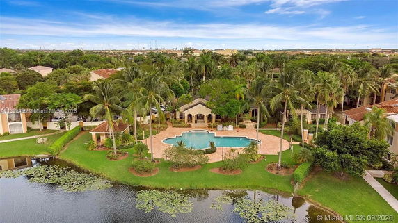 Aerial view of a swimming pool area surrounded by palm trees and greenery near a body of water.