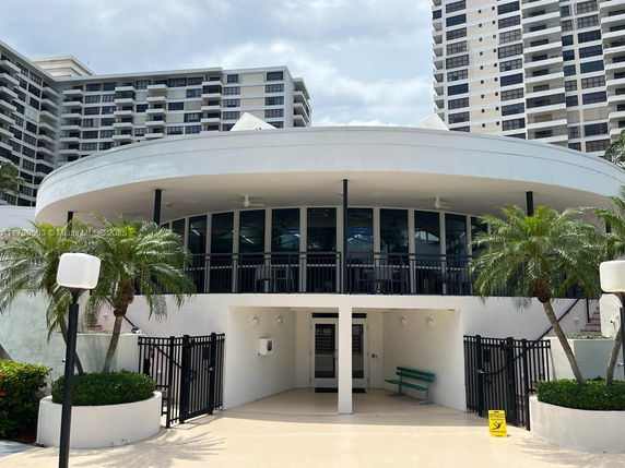 Front view of a circular building with large glass windows and a terrace, surrounded by tall residential buildings.