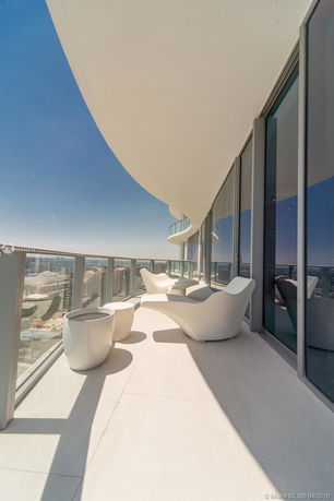 Balcony view from a high-rise apartment overlooking the city, featuring modern white chairs and glass railings.