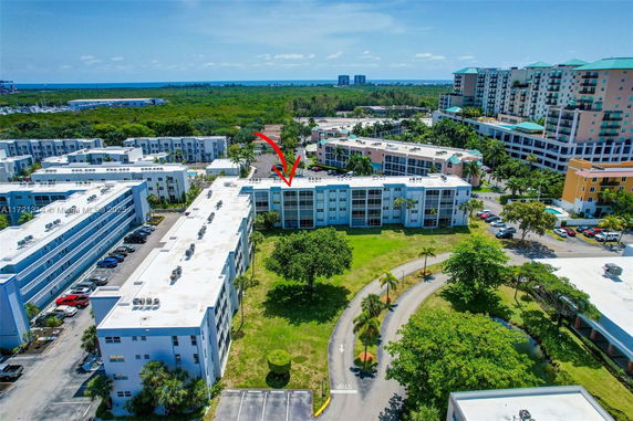 Aerial view of a complex with multiple apartment buildings and surrounding greenery, roads, and distant ocean view.