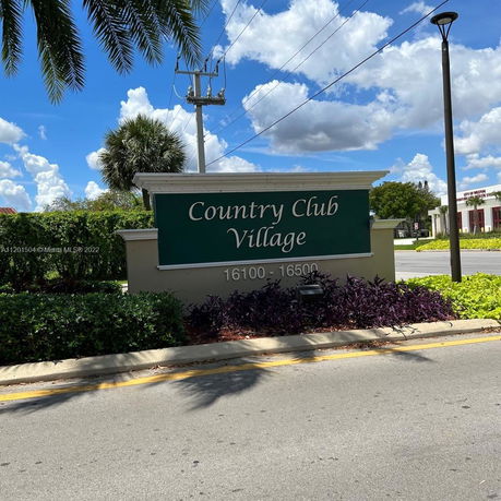 Outdoor view of a sign for Country Club Village with surrounding greenery and a clear sky.