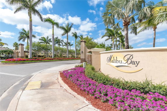 Entrance sign of Malibu Bay with palm trees and landscaped flowers.