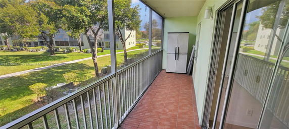 View from a screened balcony overlooking green lawn and trees with nearby buildings visible.