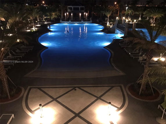 Night view of an illuminated swimming pool surrounded by palm trees and lounge chairs.