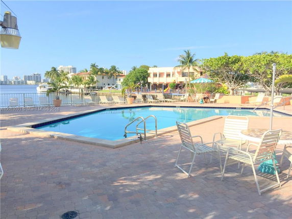 View of a building complex with a swimming pool, patio seating, and waterfront in the background.