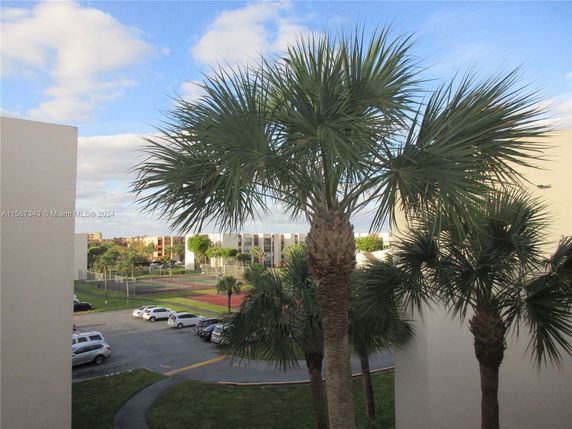 View of a tennis court and parking area with multiple parked cars. There are tall palm trees and neighboring buildings in the background.